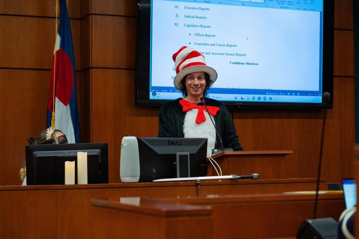 Student Body President Nick DeSalvo stands behind a podium in a wood paneled room wearing a cat costume, a large red bowtie and a red and white striped hat.