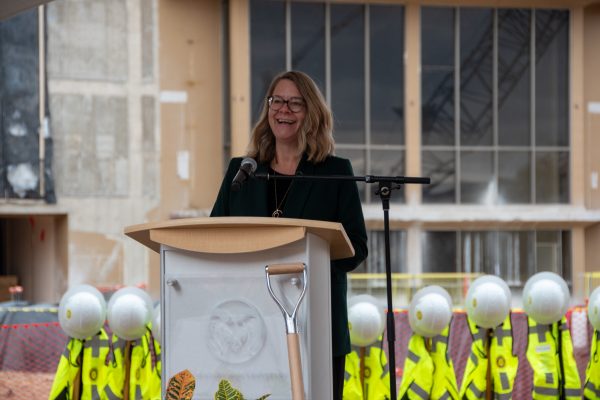 woman in black behind podium speaking to crowd with helmets and vests in background