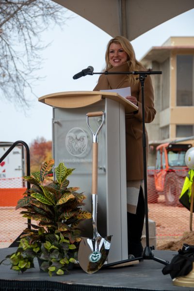 woman in brown coat speaks to a crowd behind podium