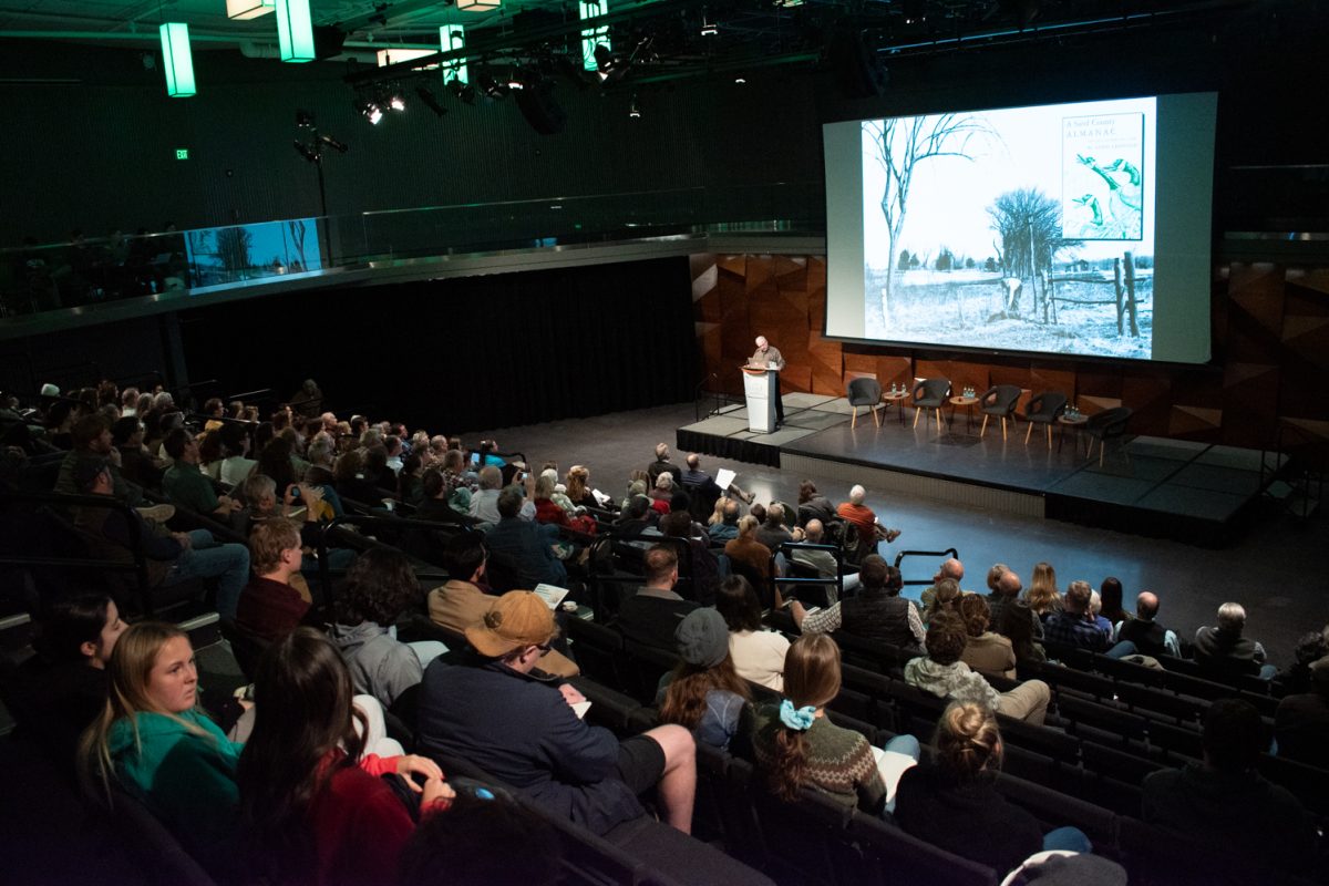 Viewed from behind, a large audience in tiered seats watches someone speak at a podium.