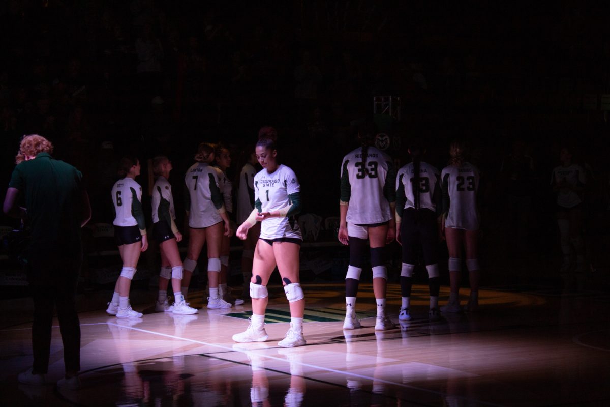 A girl wearing a white volleyball jersey with the text, "Colorado State 1" stands in a spotlight on a volleyball court.