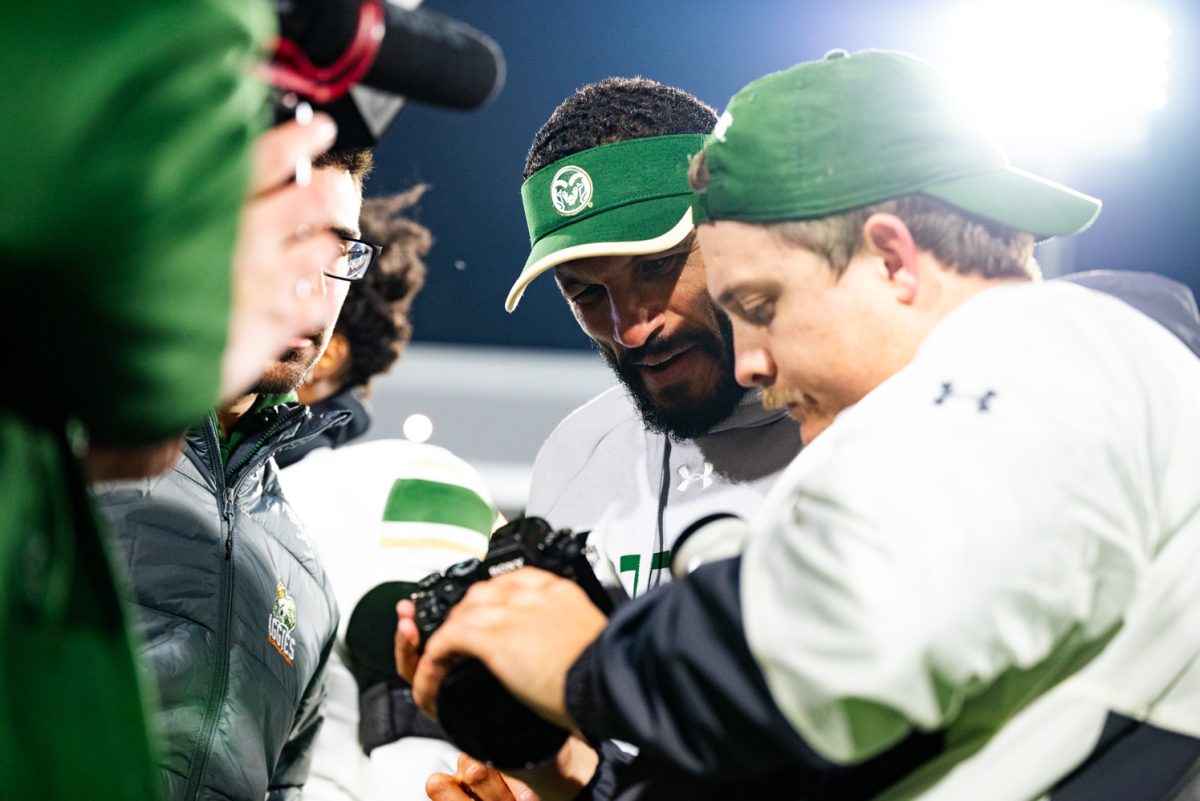 A man with a green visor looks over another man's shoulder at the camera he is holding.