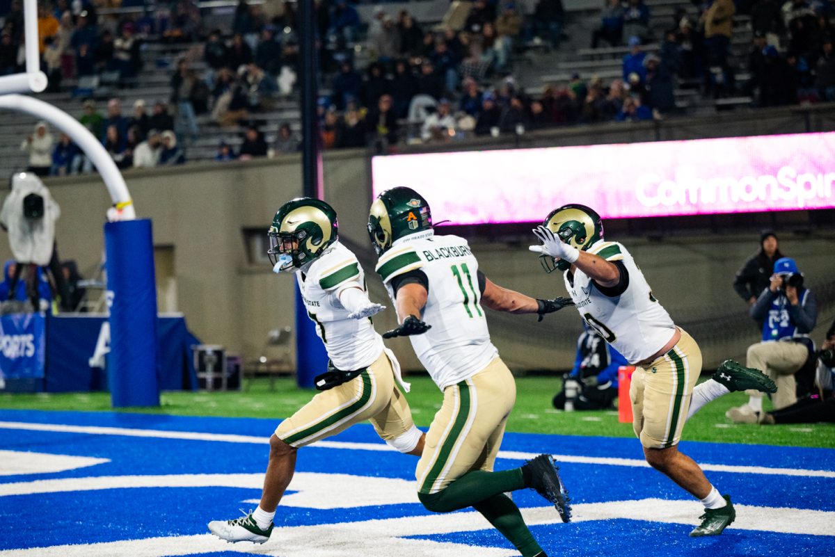 Three football players in white and green hold their arms out and run along the end zone.