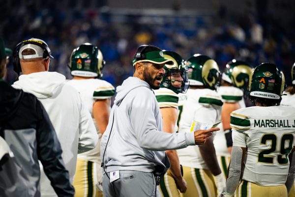 A man in a gray hoodie wearing a headset shouts and points at football players in white and green.