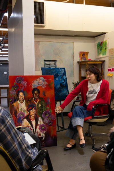 girl wearing a red sweater sits in a chair while presenting her colorful portrait painting to her classmates