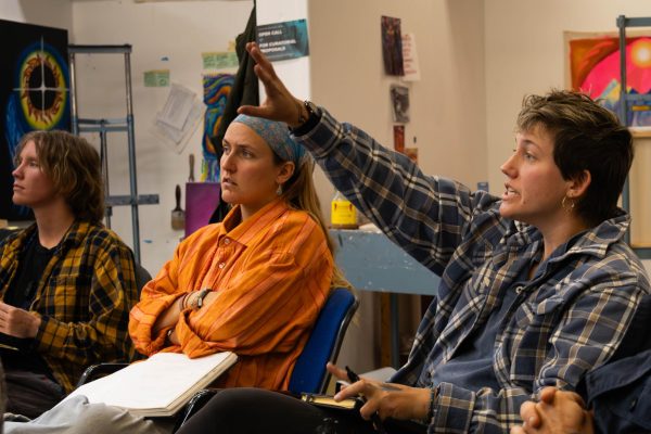 Three college students sit in a classroom art studio, wearing bright colors and flannels, discussing art.