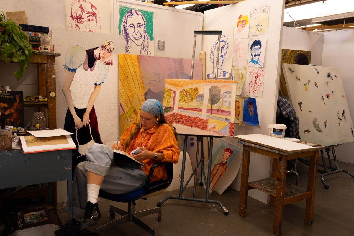 a girl in bright colors sits in college class studio working. Drawing in her sketch book, surrounded by her colorful portrait paintings.