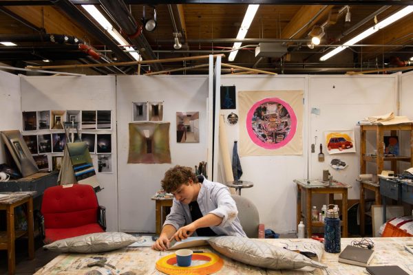 person works on a multimedia art project during their painting capstone class. With art hanging on the walls behind them, the person works on attaching wing shaped pillows to a painting of a car.