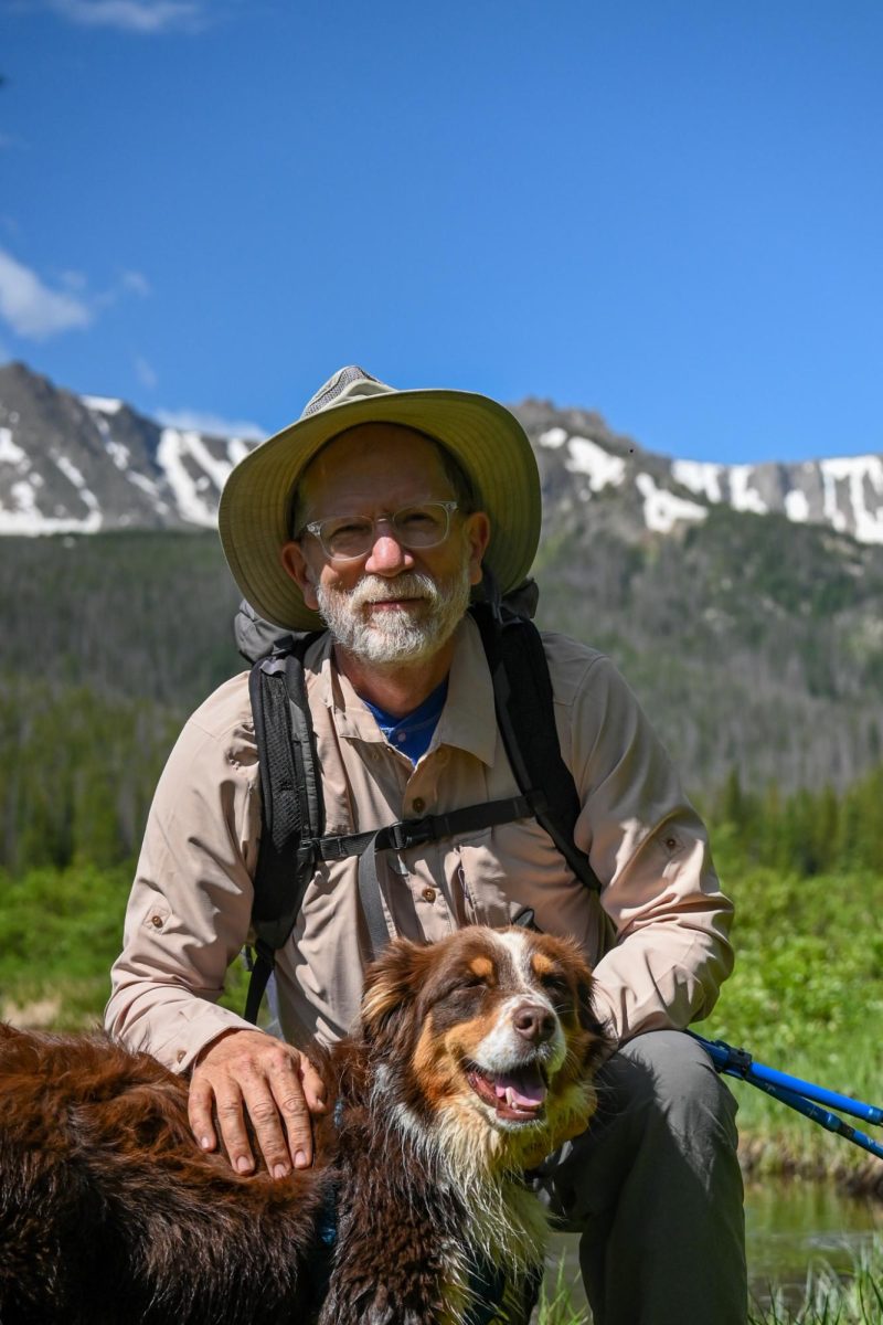 An older man in hiking clothes kneeling next to his brown dog.