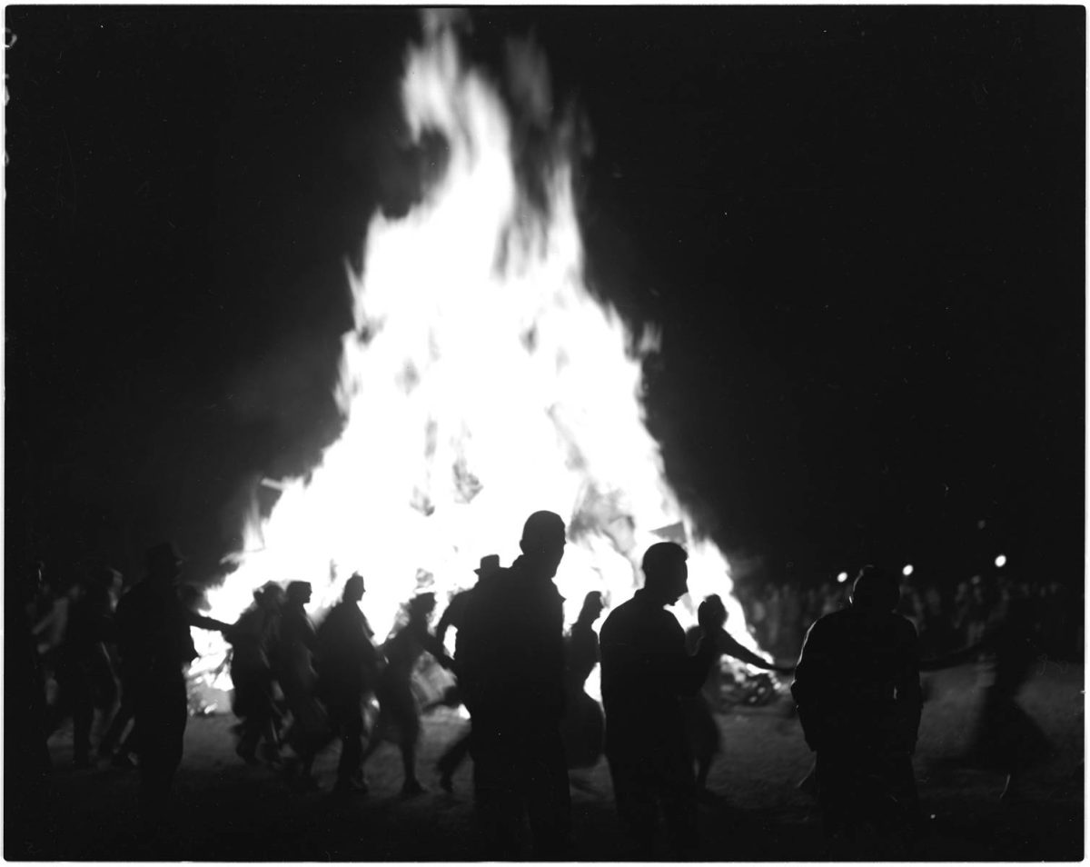 A black and white photo of a bonfire with people dancing around it.