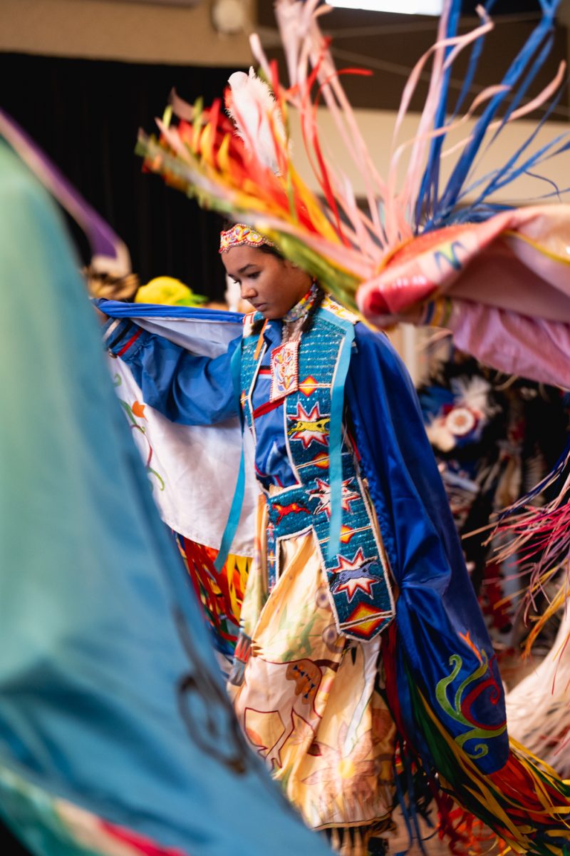 A dancer in traditional Native American regalia with an array of colors dances in a ballroom.