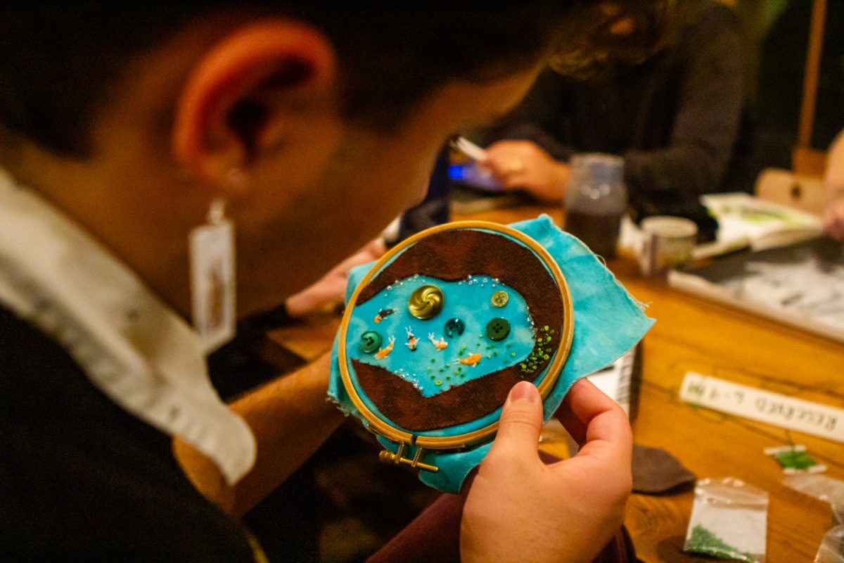 A man embroiders a piece of blue fabric with beads, buttons, and felt.