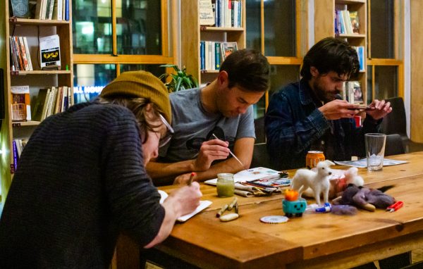 Three men sit at the end of a wooden table and draw in a bookshop.