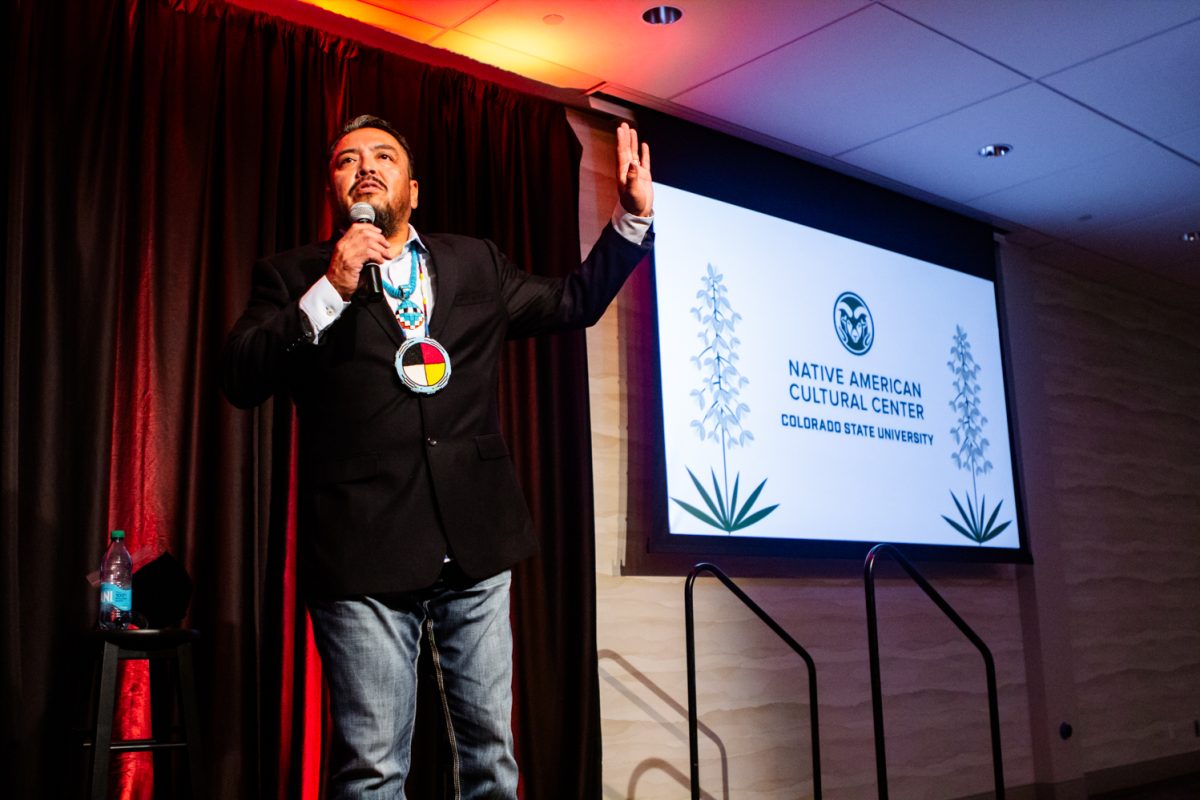 A man speaks on a small stage, while a presentation slide reading "Native American Cultural Center Colorado State University" is projected behind him.