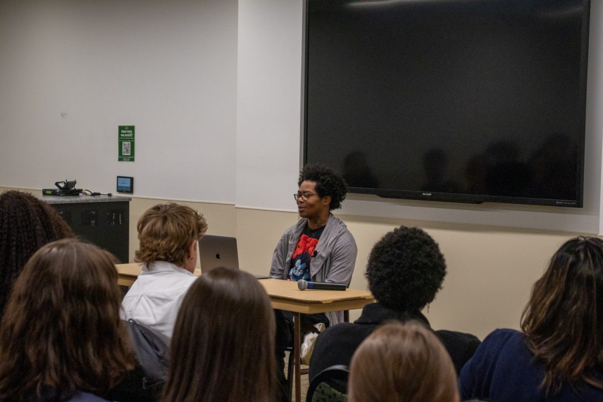 A person sits at a table looking at a laptop speaking to a room of people.
