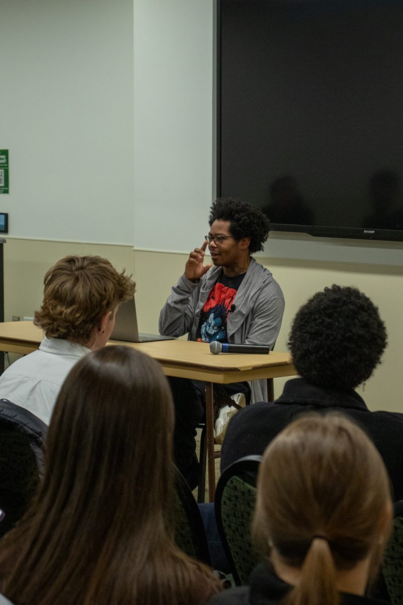 A person sits at a table looking at a laptop speaking to a room of people.