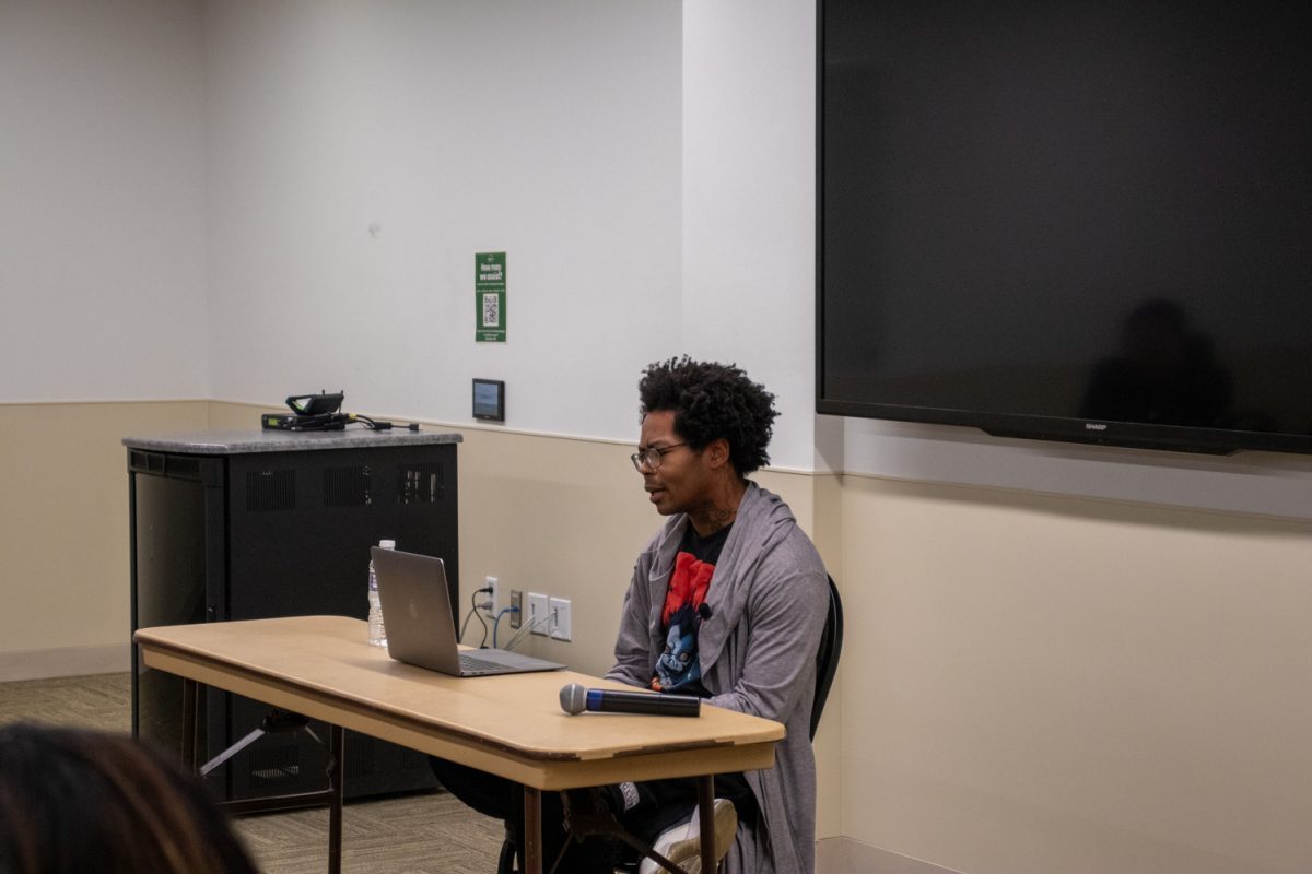A person sits at a table looking at a laptop speaking to a room of people.