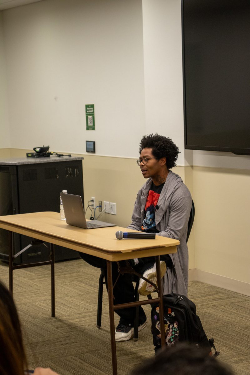 A person sits at a table looking at a laptop speaking to a room of people.