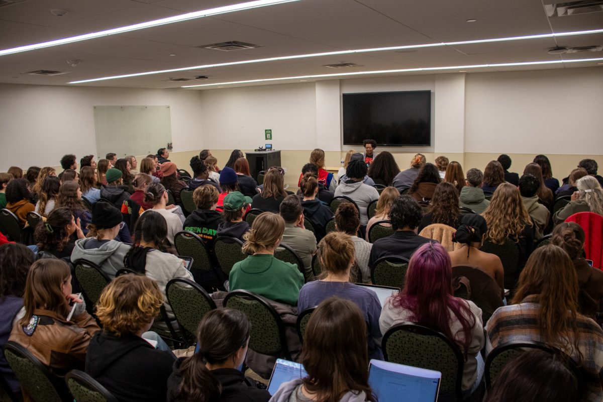 A person sits at a table looking at a laptop speaking to a room of people.