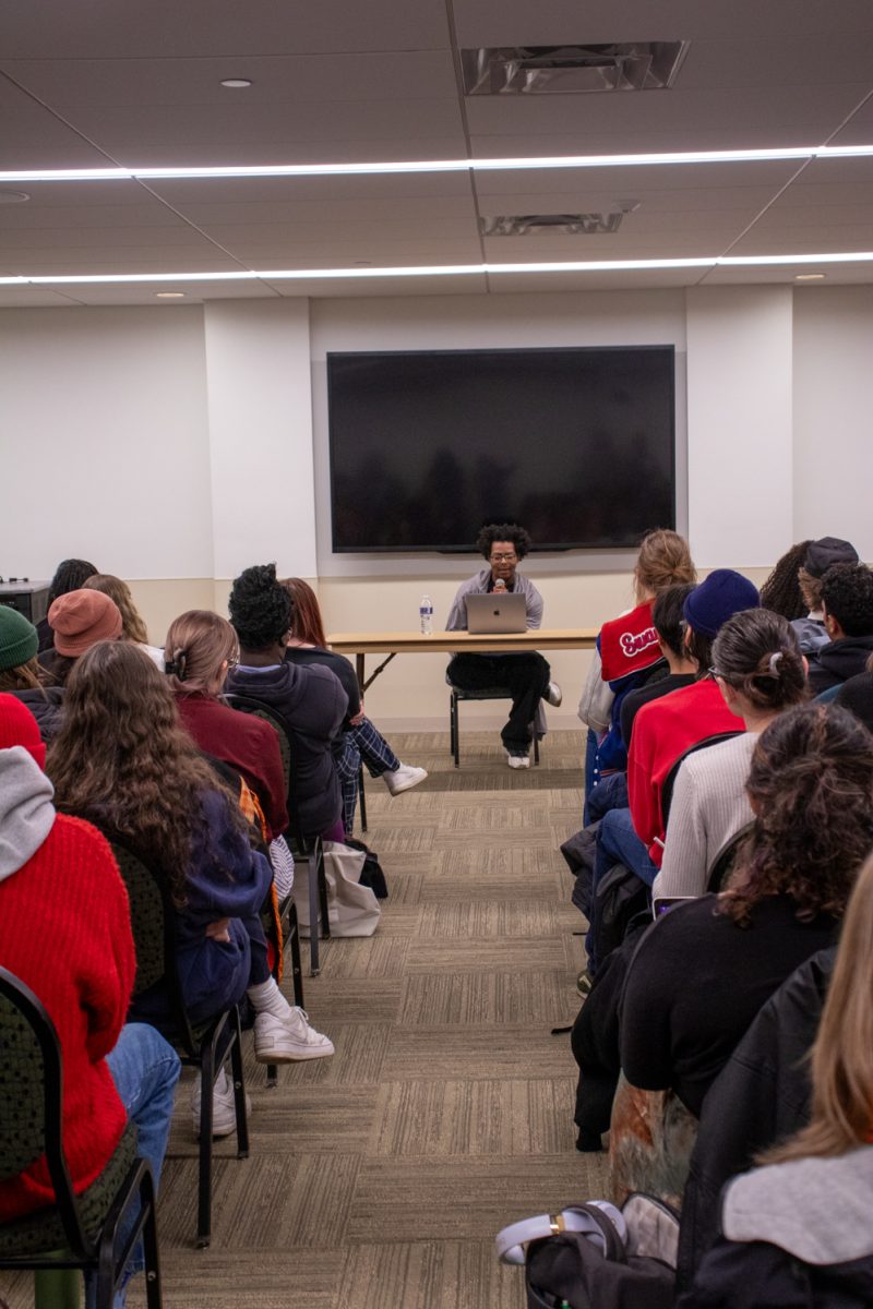 A person sits at a table looking at a laptop speaking to a room of people.