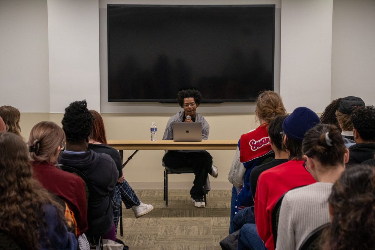 A person sits at a table looking at a laptop speaking to a room of people.
