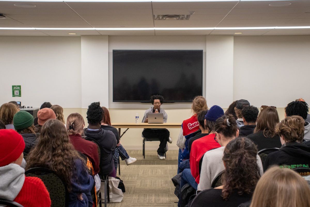 A person sits at a table looking at a laptop speaking to a room of people.