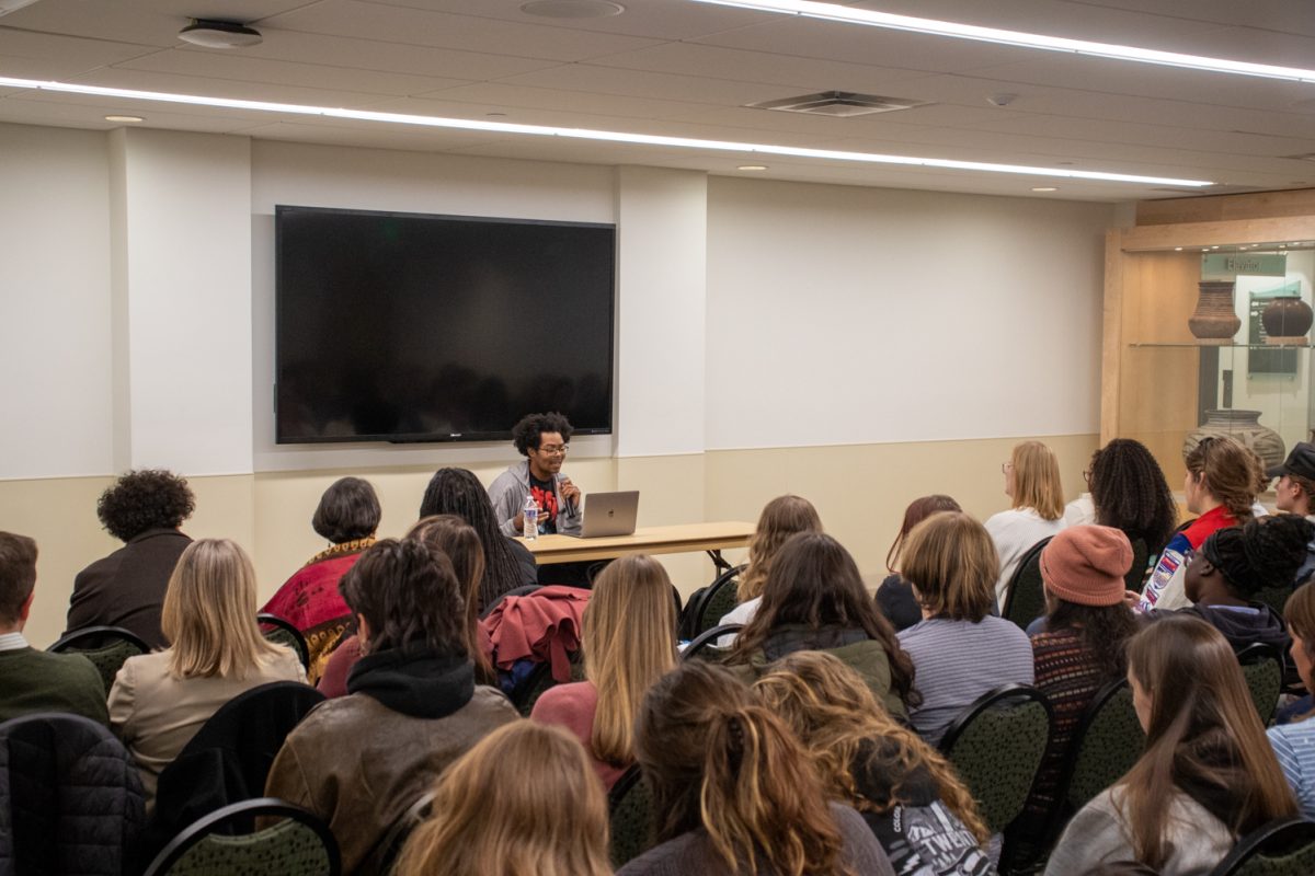 A person sits at a table looking at a laptop speaking to a room of people.