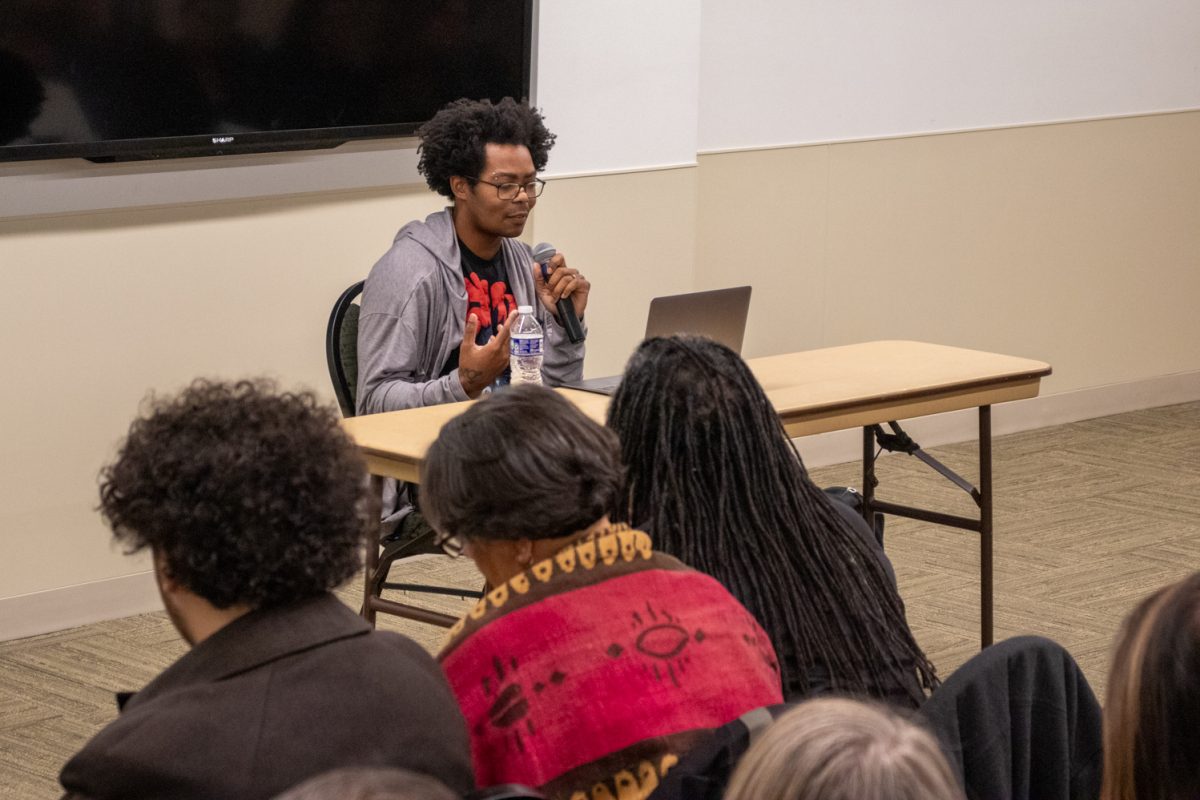 A person sits at a table looking at a laptop speaking to a room of people.