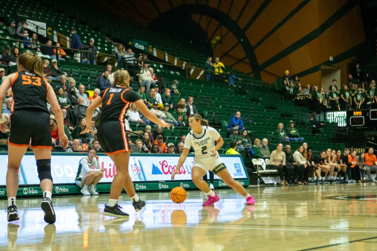 A woman in a white jersey bounces an orange ball around women in black jerseys at a sporting event.
