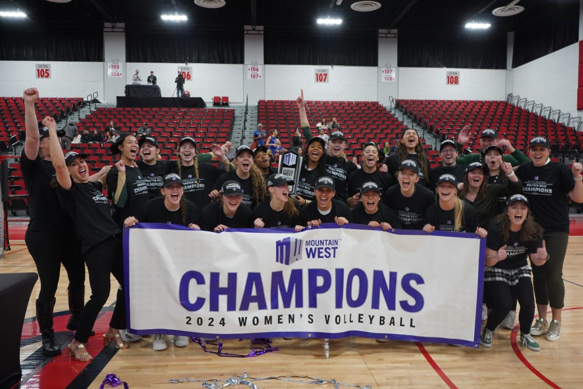 A group of smiling woman volleyball players hold a banner reading "Champions" in a gymnasium.