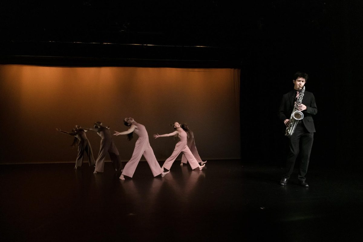 Five dancers in pink jumpsuits dance on a stage. In the foreground, a man in a black suit plays a silver saxophone.