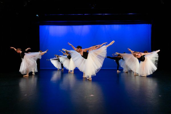 Dancers in white skirts and black tops pose on stage during a performance.