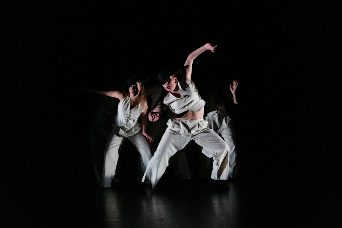 Three dancers in white on a completely dark stage.