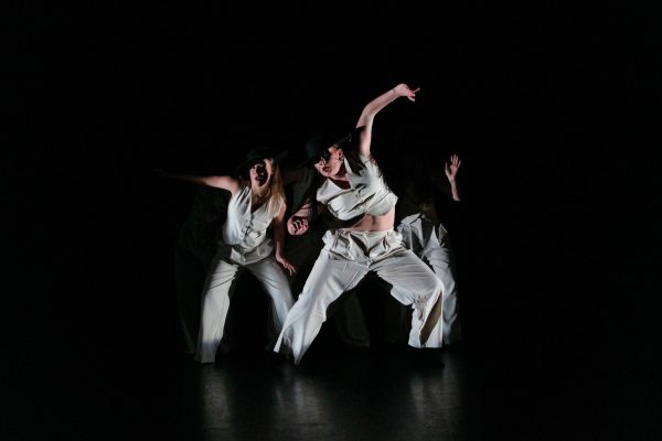 Three dancers in white on a completely dark stage.