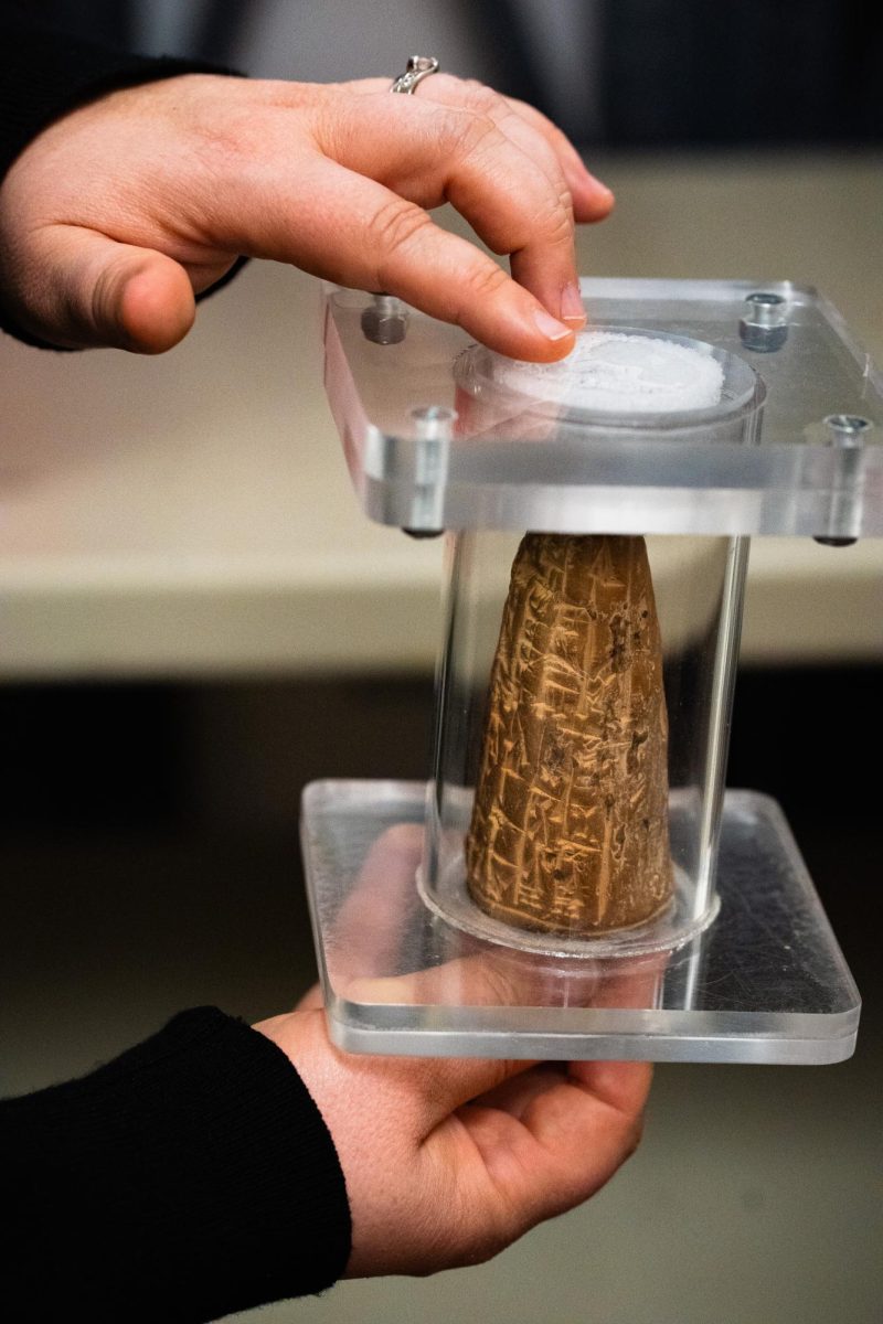 A close up photo of two hands hold an artifact in a protective clear casing. There is cuneiform on the conical artifact.