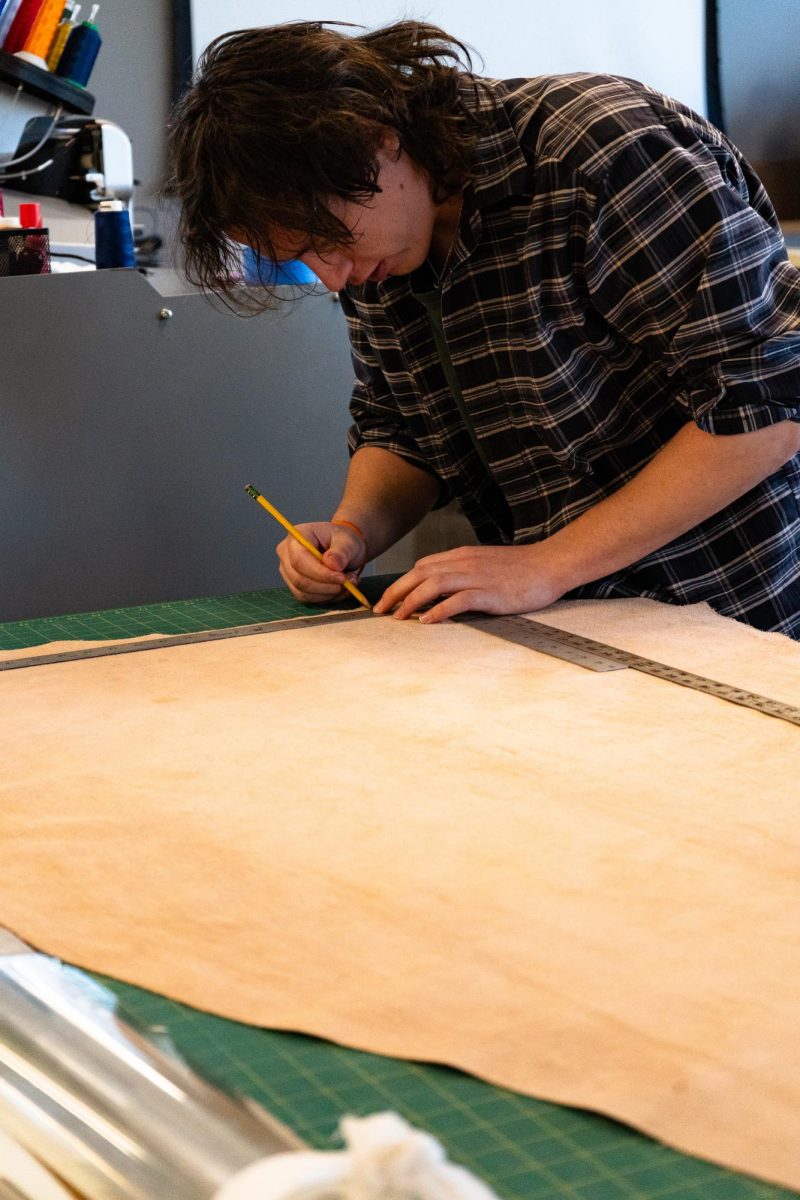 A student leans over a large piece of tan leather on a cutting table. Two metal rulers are on the leather as the student uses a pencil to trace lines onto the material.