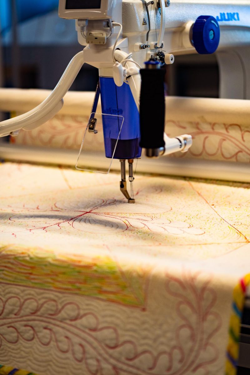 A machine with thread fed through it and a needle for embroidery rests on a partially completed quilt.