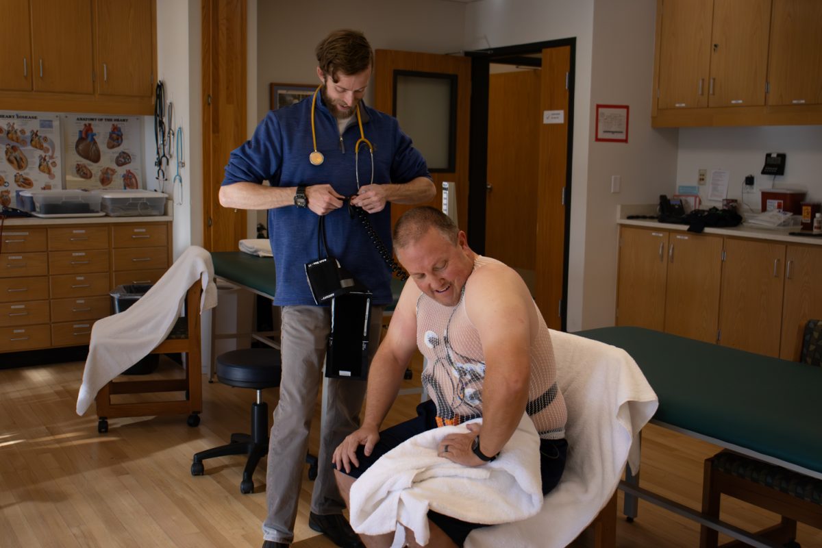 In a medical room, a young doctor wearing blue shirt prepares to take the heart rate of a middle aged male who is getting his heart work done