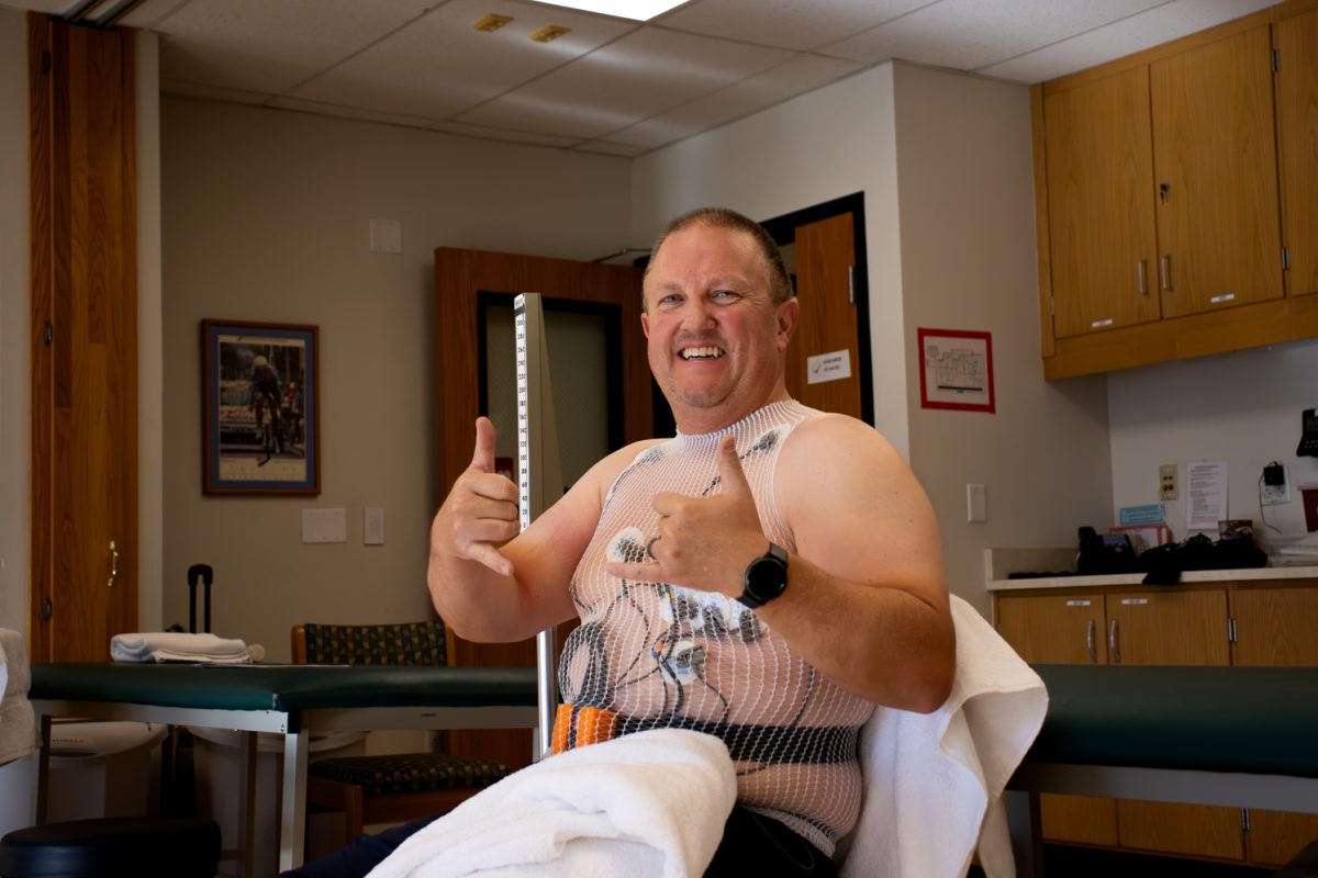 middle aged man gets attached to wires with a fish net shirt to hold the wires in place gets his heart work done and hold up a Shaka hand gesture with both hands as he smiles at the camera