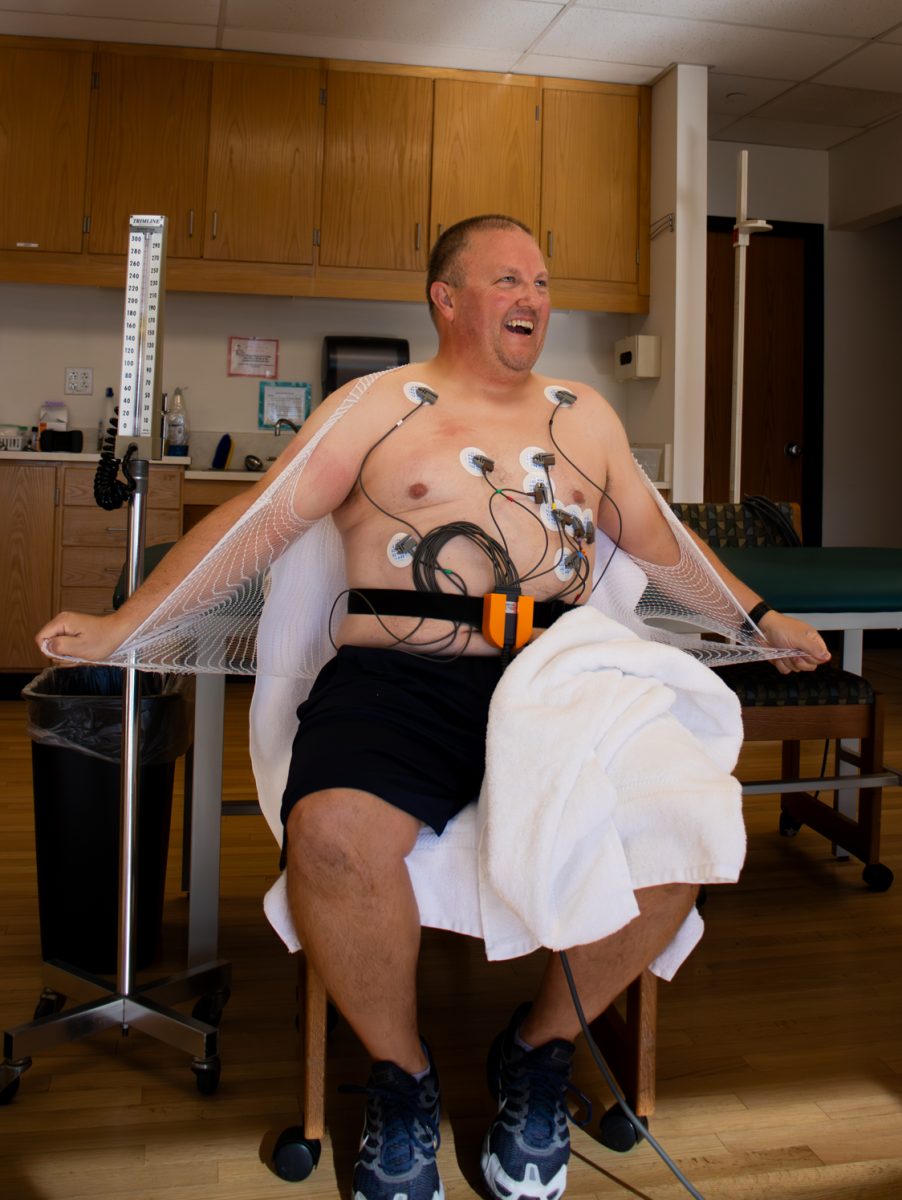 local male firefighter gets his heart rate monitored. Sitting down and attached to multiple wires along his chest to monitor his heart and he sits in a chair and rips off the mesh fishnet shirt given to him to hold to wires in place during testing