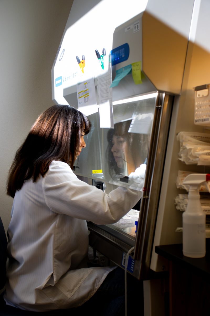 Female student wearing a white lab coat with dark brown hair works in a lab room. Her afce reflects off of the protective glass, creating a reflection looking back at herself