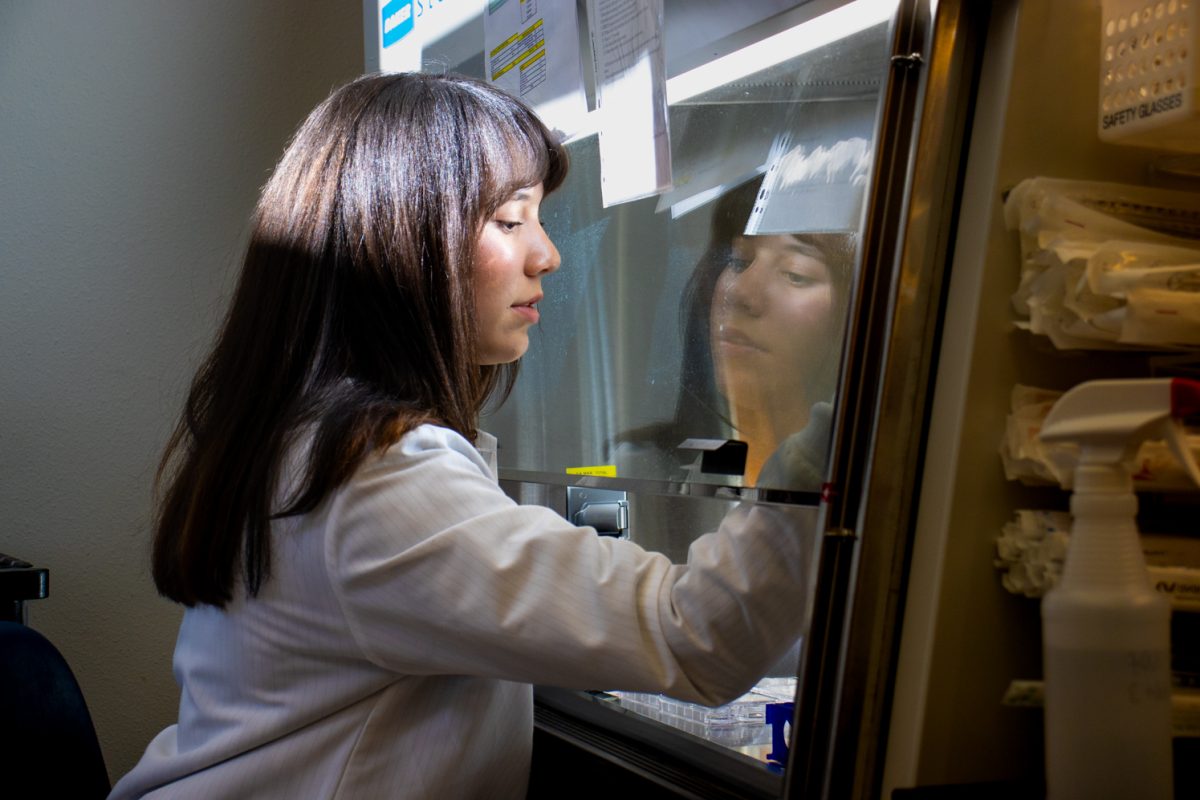 Female student wearing a white lab coat with dark brown hair works in a lab room. Her afce reflects off of the protective glass, creating a reflection looking back at herself