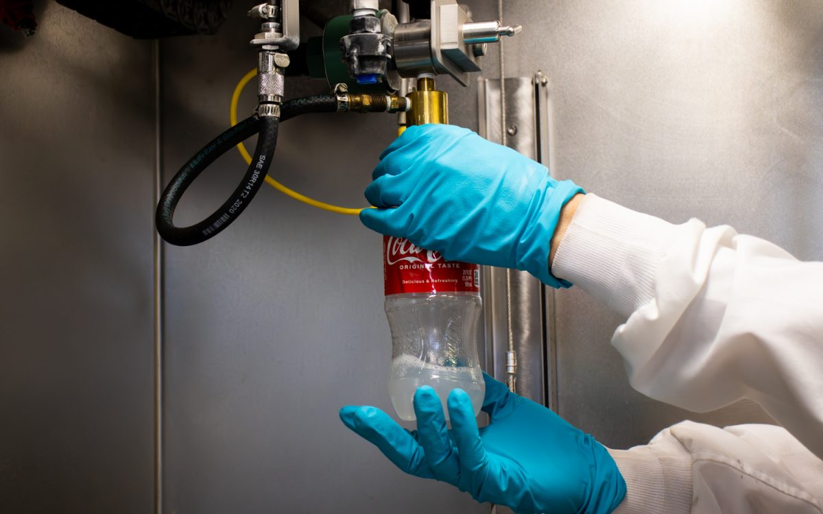 a pair of hands wearing bright blue protective gloves puts an old coca-cola water bottle that contains a liquid substance into machine to be used for science