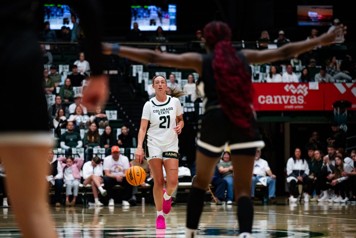 A female basketball player in white and green calmly dribbles a basketball towards a player in black and white with her arms spread out.