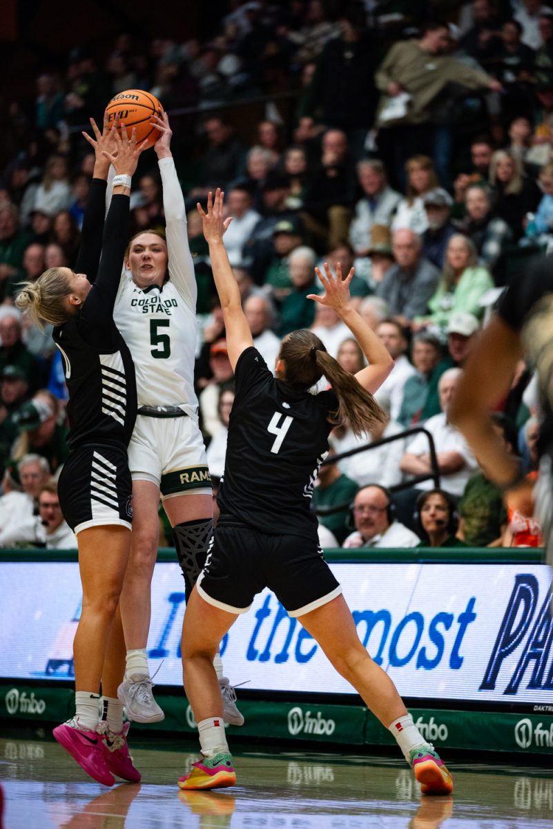 A female basketball player in white and green jumps up with a ball while two players in black and white try to stop her.