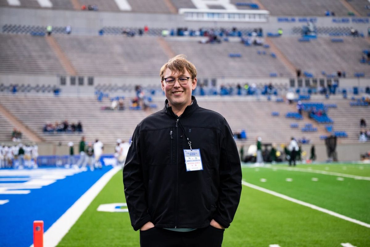 A man in a black jacket with a "Football Media" press pass poses for a photo on the sidelines of a football field.