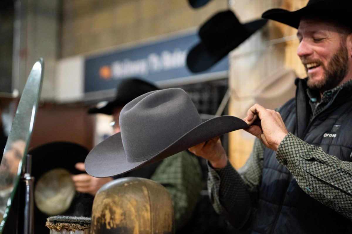 A man with a green shirt, a black vest and a black cowboy hat steams a cowboy hat for a customer.