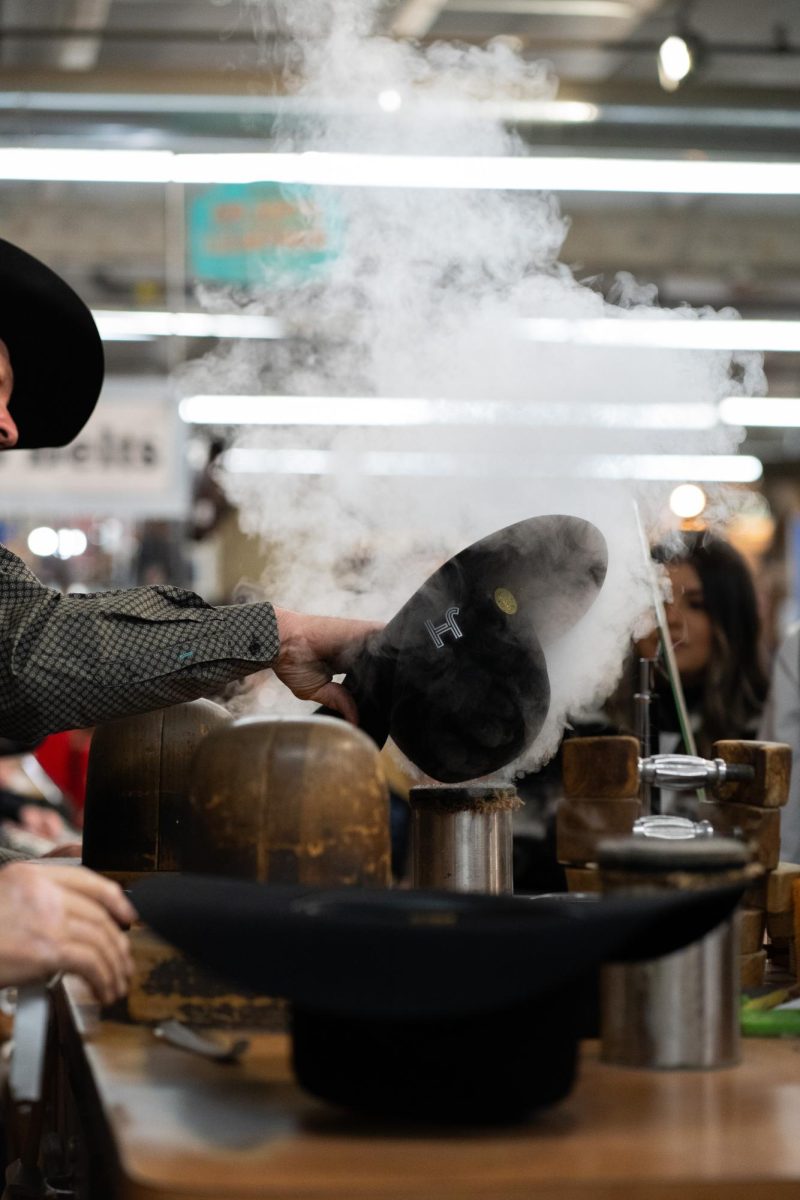 A man with a green shirt, a black vest and a black cowboy hat steams a cowboy hat for a customer.