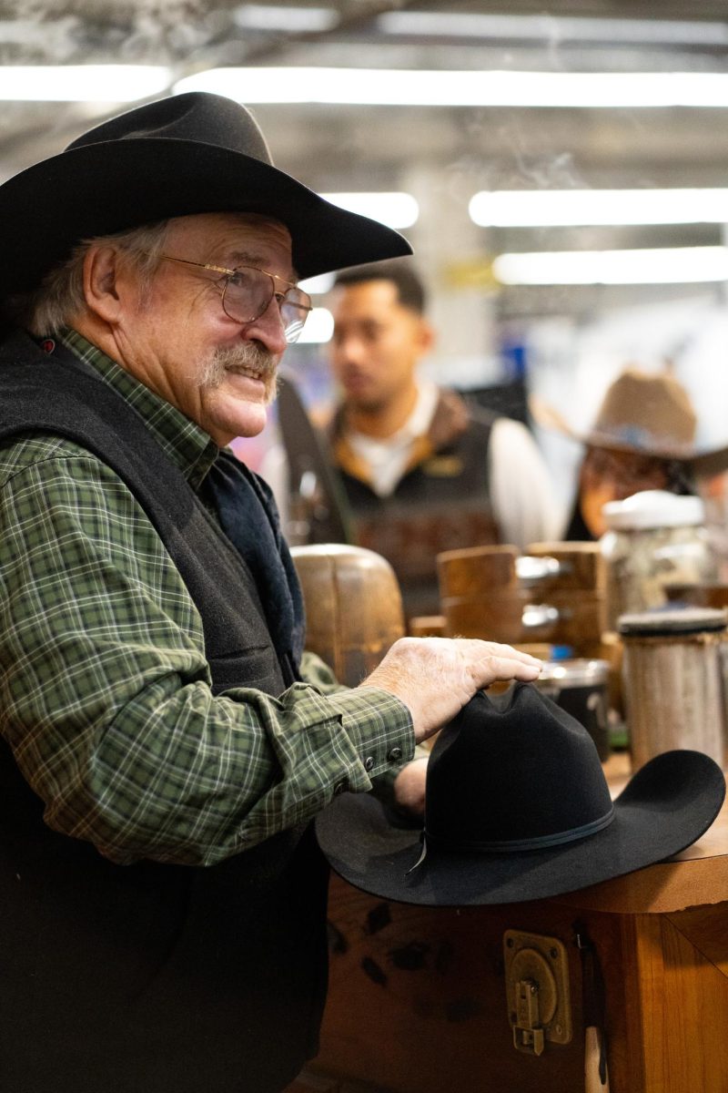 A man with a green shirt, a black vest and a black cowboy hat shapes a customer's black hat.