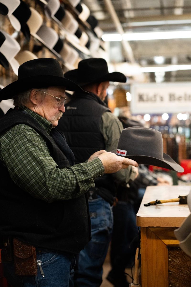 A man with a green shirt, a black vest and a black cowboy hat shapes a hat for a customer.